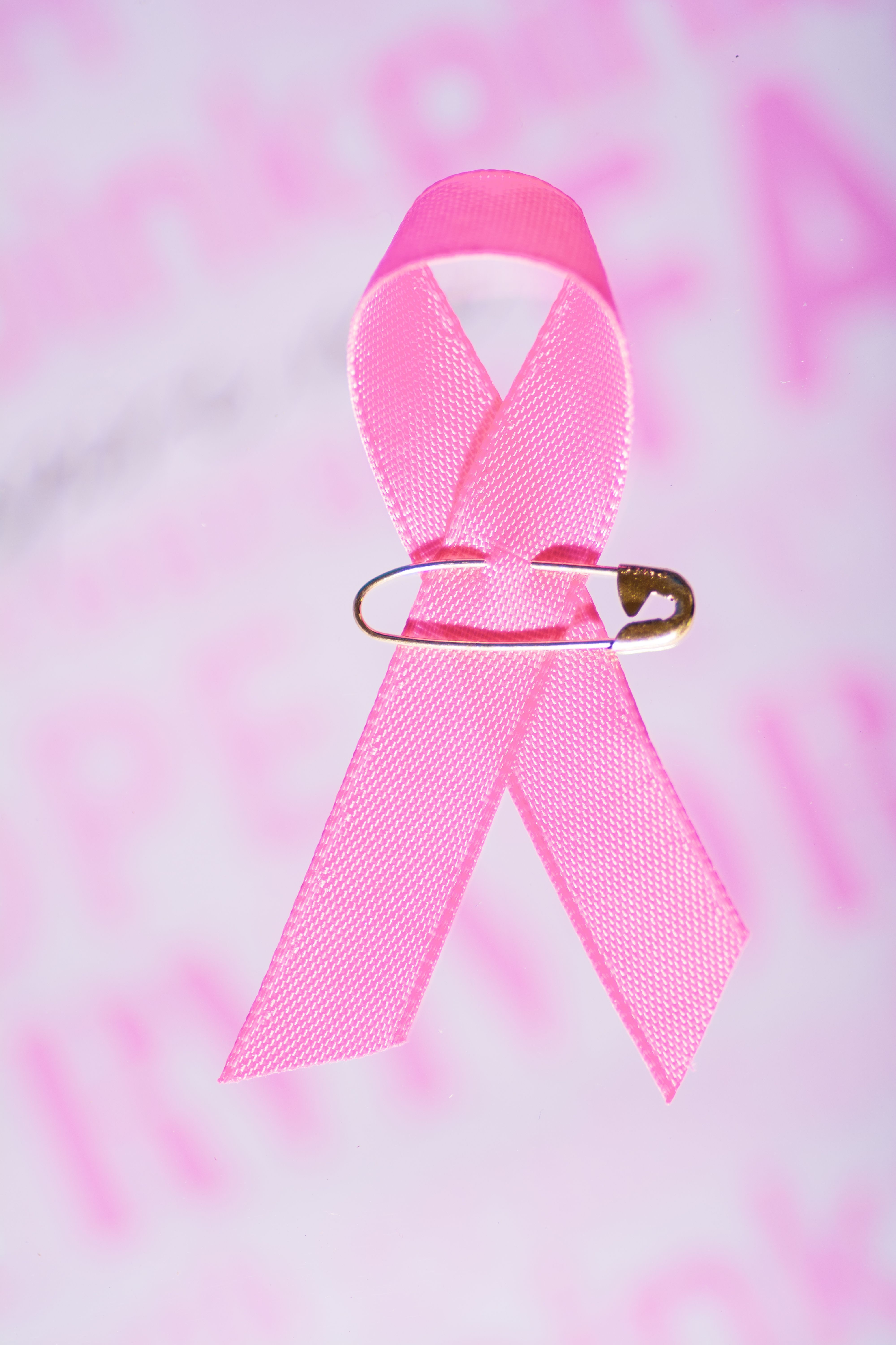 Pink breast cancer awareness ribbon held with a silver safety pin against a soft pink background
